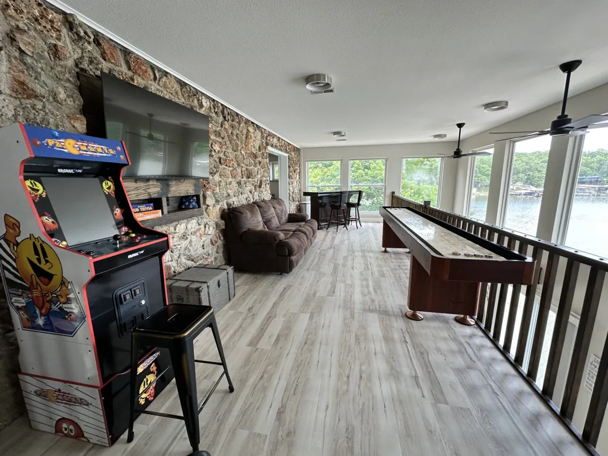 An airy, sunlit game room featuring a classic arcade machine, shuffleboard table, and comfortable seating. Large windows provide views of the outdoors, and the setup is clearly geared toward fun and group activities.