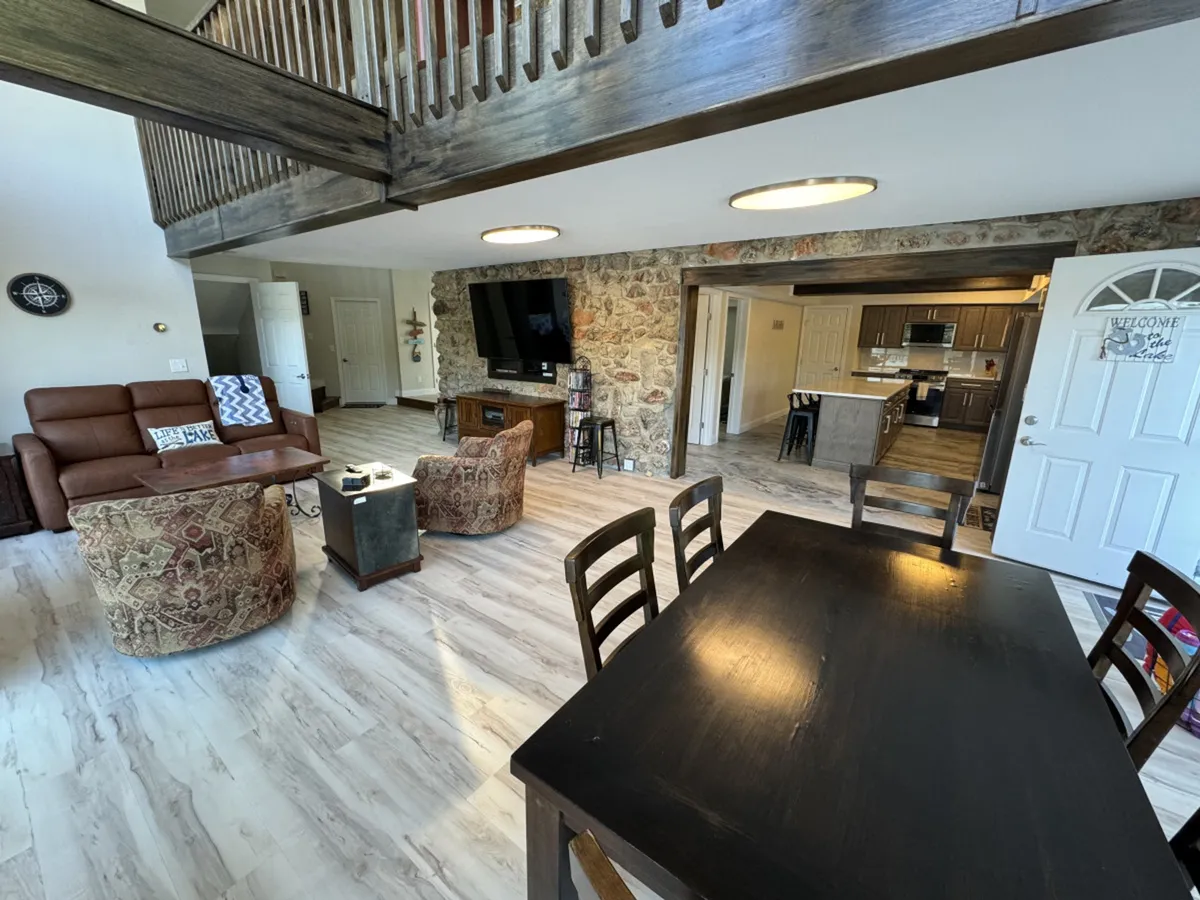 A spacious open-plan living and dining area featuring a striking stone wall, a large dining table ready for group meals, a comfortable sofa set, and a flat-screen television. The arrangement is optimized for socializing, relaxing, and entertaining, capturing the essence of communal family and group gatherings.