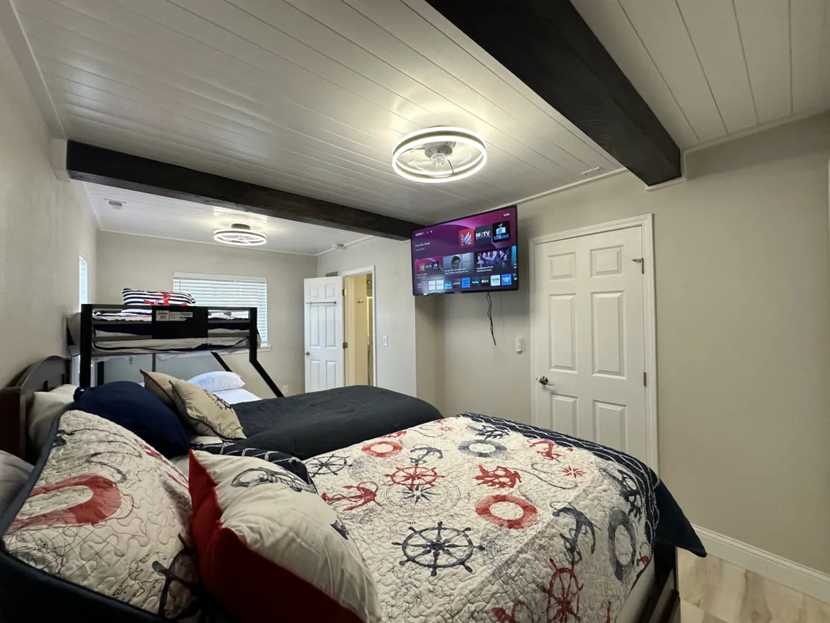 A functional and comfortable bedroom setup featuring a main bed, a bunk bed, a wall-mounted TV, and decorative bedding. The layout encourages relaxation and family time, whether watching a movie or winding down before bed.