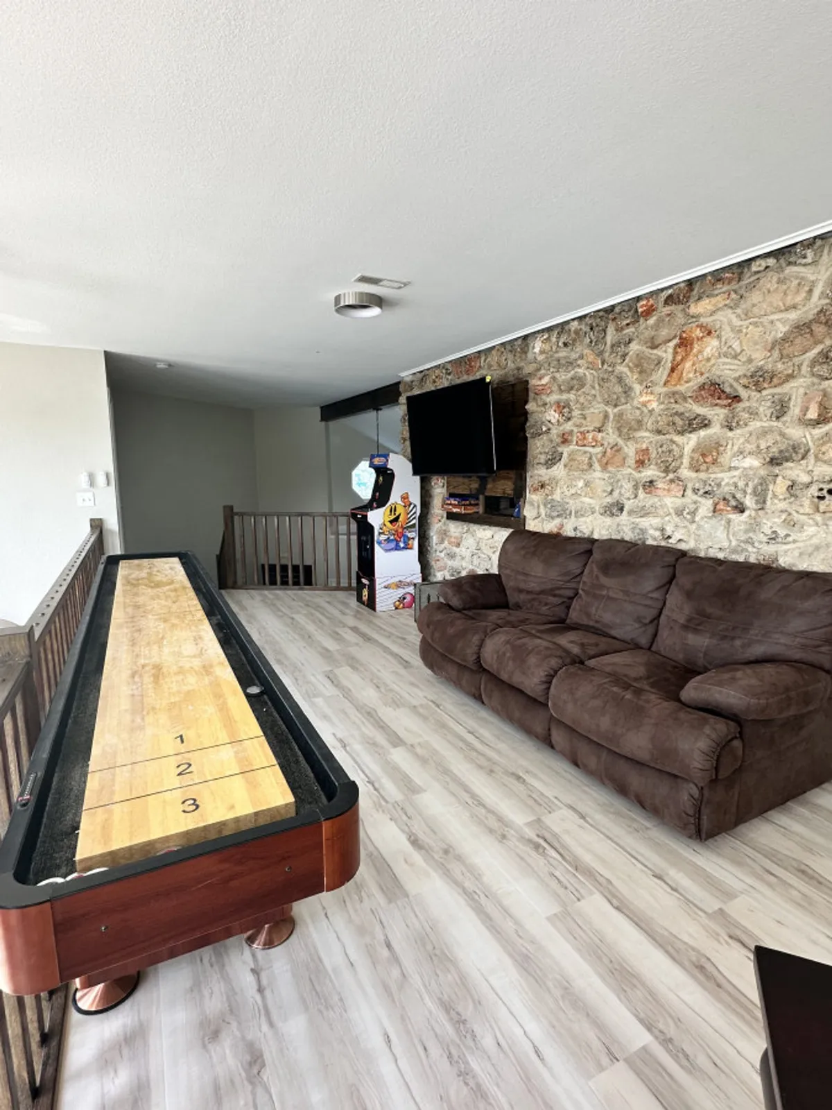 A shuffleboard table is the main focus, accompanied by a brown couch, a stone wall, and a television. This space looks ready for both friendly competition and relaxed movie watching, highlighting versatility.