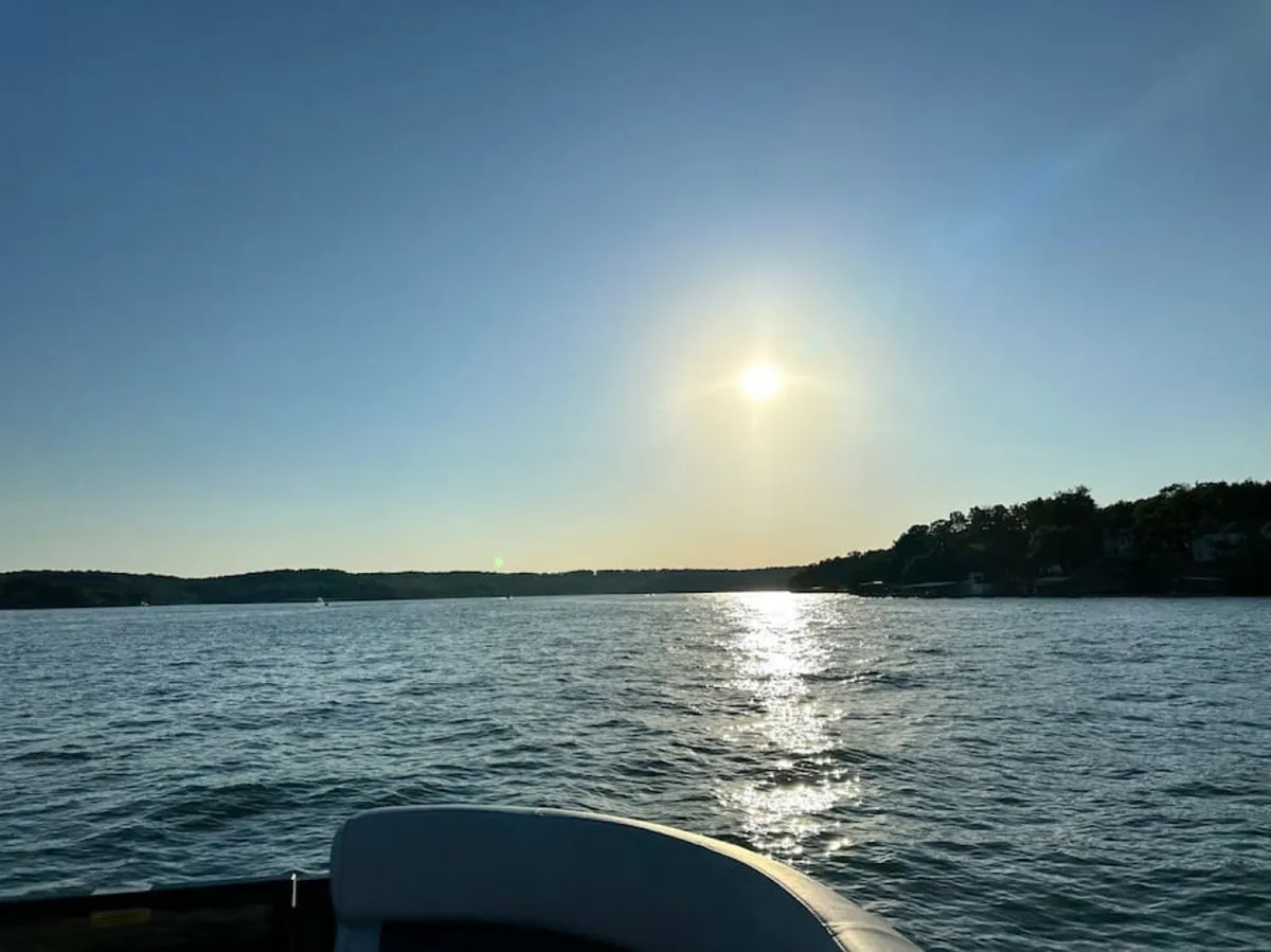 The image showcases a breathtaking, serene view across the sparkling surface of Lake of the Ozarks. The sun begins to set, reflecting golden light on the water, with the distant shoreline visible. The scene evokes relaxation and the allure of lake activities such as boating and fishing.