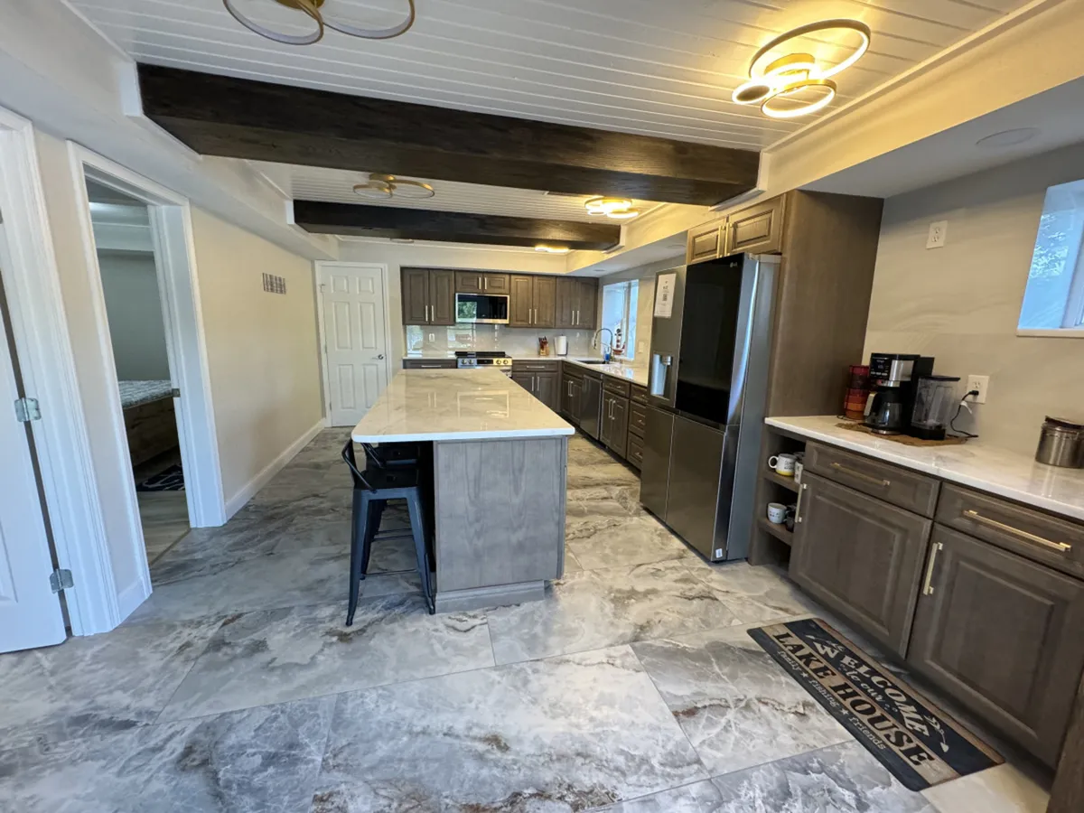A spacious kitchen with an island featuring bar seating, stainless steel appliances, and beamed ceilings. The room is filled with natural light and designed as a central hub for cooking, socializing, and family gatherings.