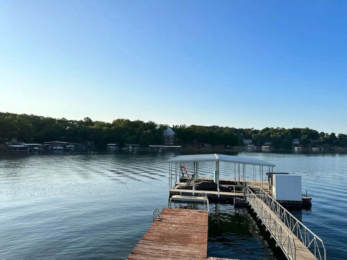 A wide exterior view of the dock stretching over the calm lake, surrounded by a lush tree line and houses in the background. The soft lighting suggests either an early morning or evening, highlighting the peacefulness of the setting.