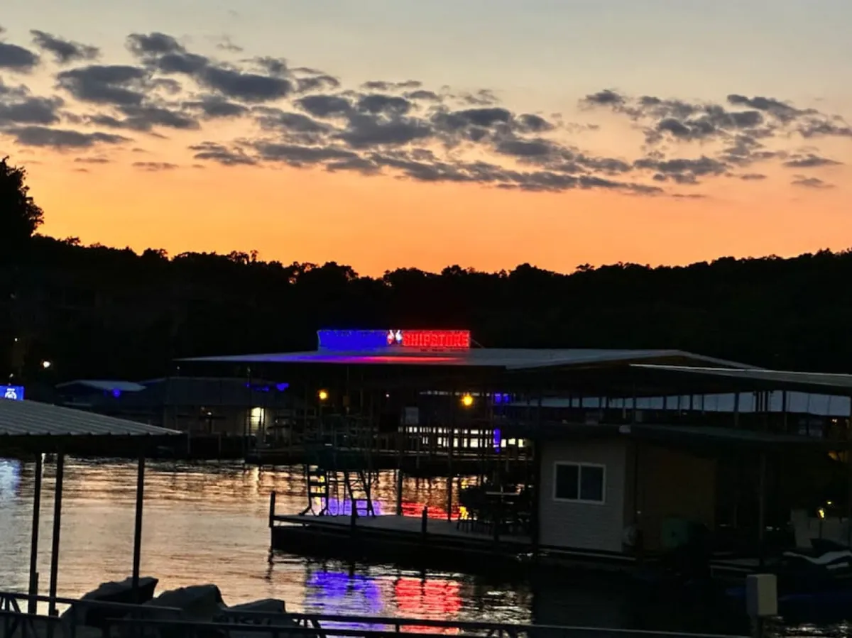 An exterior shot of a marina at sunset, with an illuminated building reflecting on the water. The setting sun paints the sky with a warm glow, suggesting a lively social atmosphere for dining, listening to music, or enjoying a drink after a day on the water.
