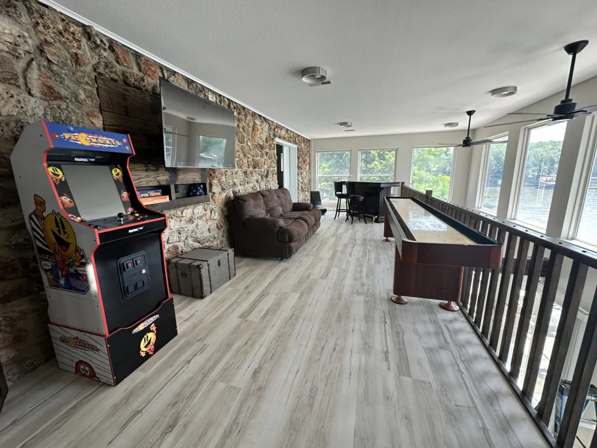 The scene includes an arcade machine, stylish sofa, bar table, large windows, and a shuffleboard table. The space feels bright and sociable, perfect for groups, and invites both play and relaxation.