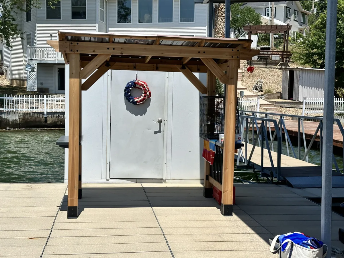 A festive wooden pergola with a patriotic wreath and a white door sits at the edge of a patio, ideal for lakeside gatherings, cookouts, or relaxing in the shade with friends and family. The space suggests outdoor entertaining with welcoming and cheerful details.