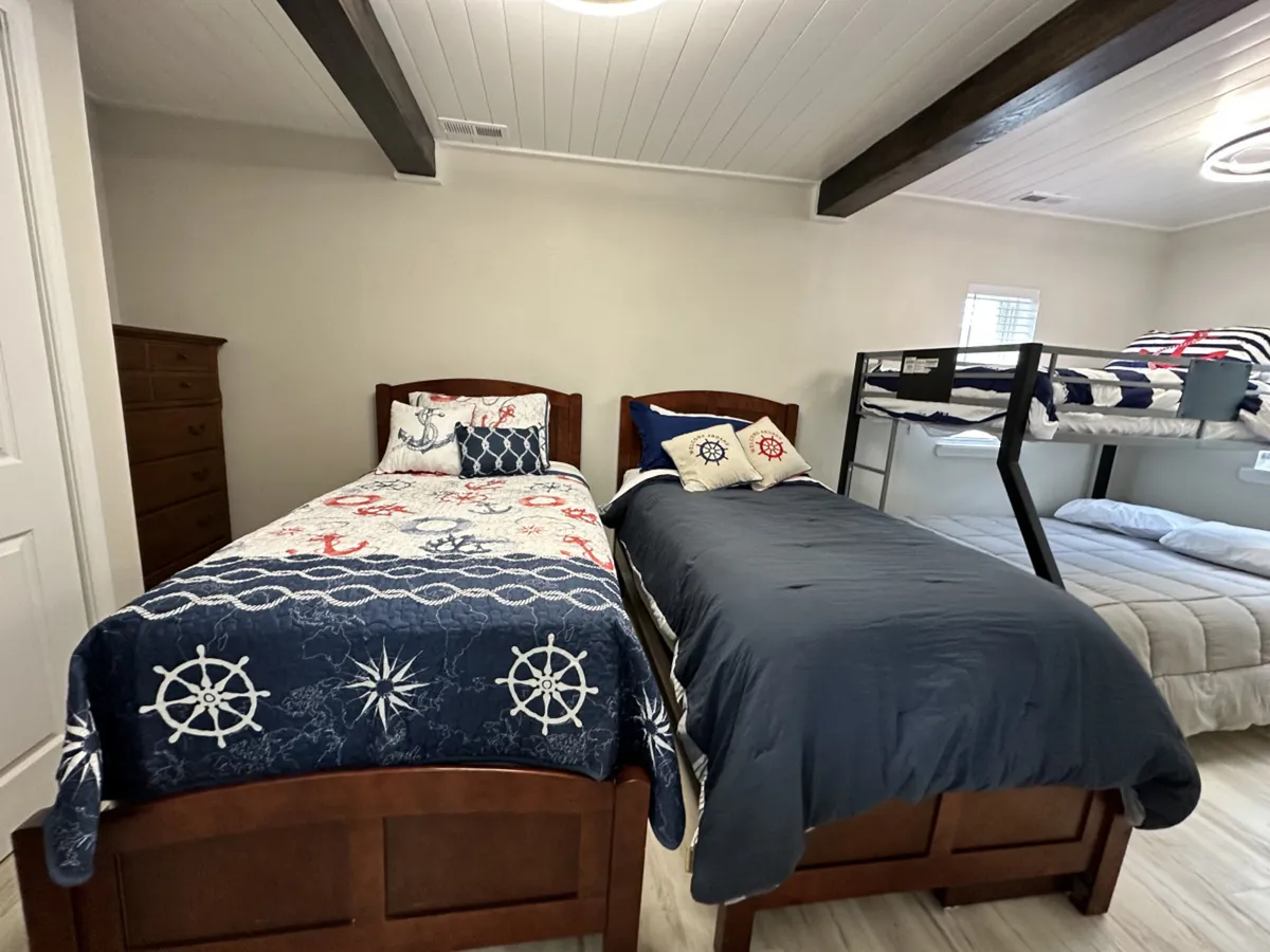 A cozy bedroom designed for families or children, featuring two beds with nautical-themed bedding and a bunk bed in the corner. A wooden dresser provides additional storage, and the space is decorated in a playful yet inviting style.