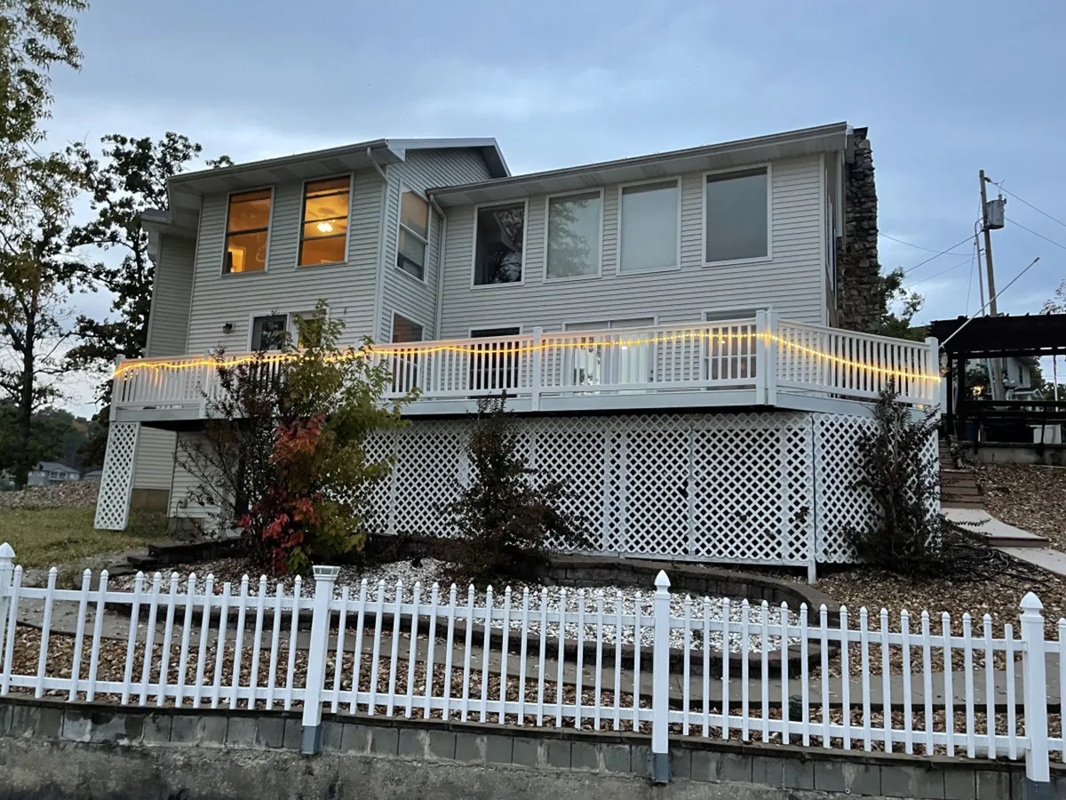 This exterior shot features a welcoming deck illuminated by string lights, a white picket fence, and expansive windows that suggest spacious interiors. The scene feels inviting, with the deck clearly set up for gatherings, relaxation, and evening enjoyment under the stars.
