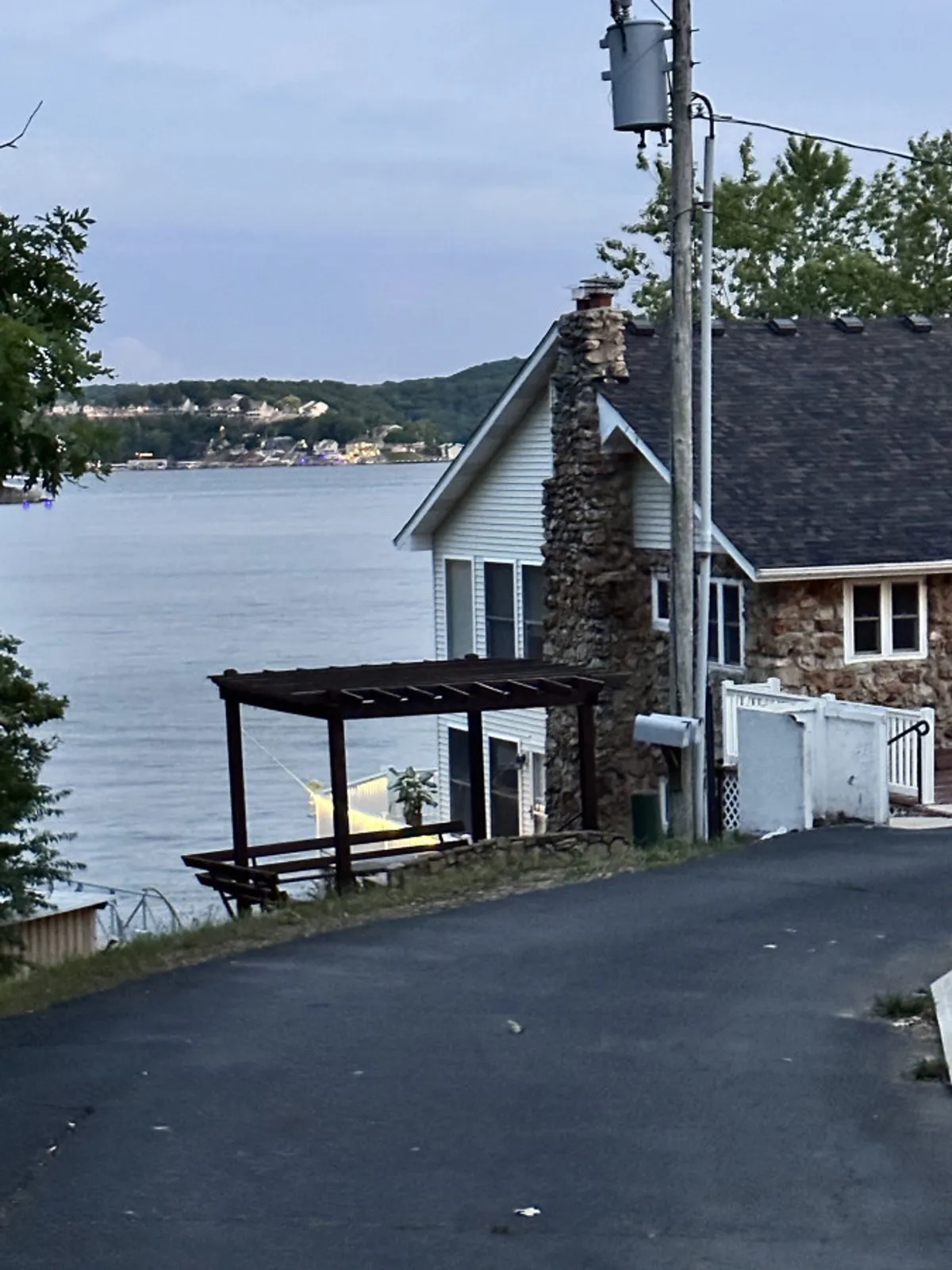 This image showcases a serene lake view from the property, including a stone wall, a wooden pergola, and the tranquil water beyond. The setting is ideal for relaxing by the lake, hosting gatherings, or simply enjoying the peaceful surroundings.
