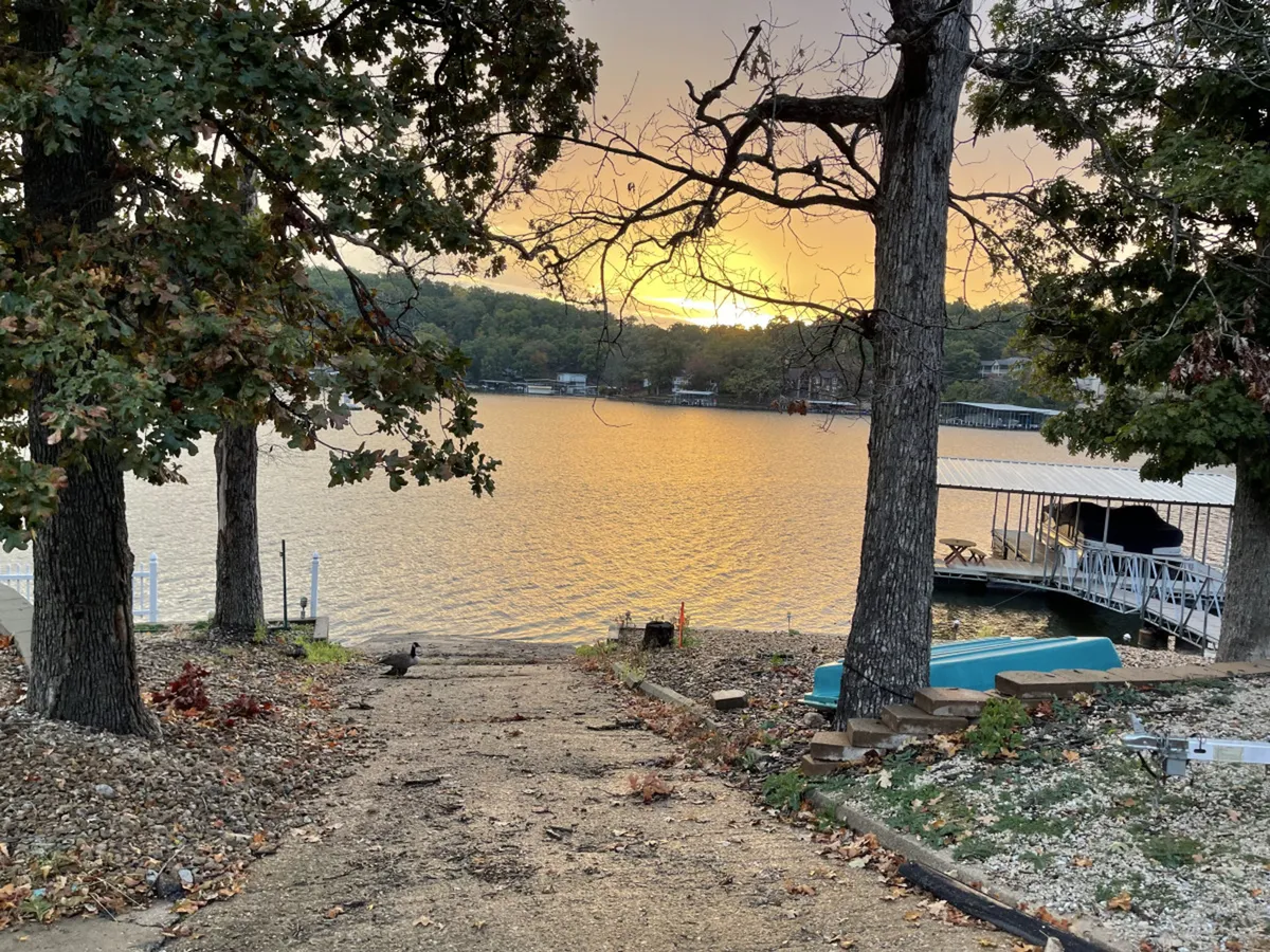 A picturesque sunset over the water, with a path winding down to the lake, docks visible in the distance, and trees framing the scene. The mood is tranquil, evoking evenings spent fishing, boating, or watching the sunset by the water's edge.